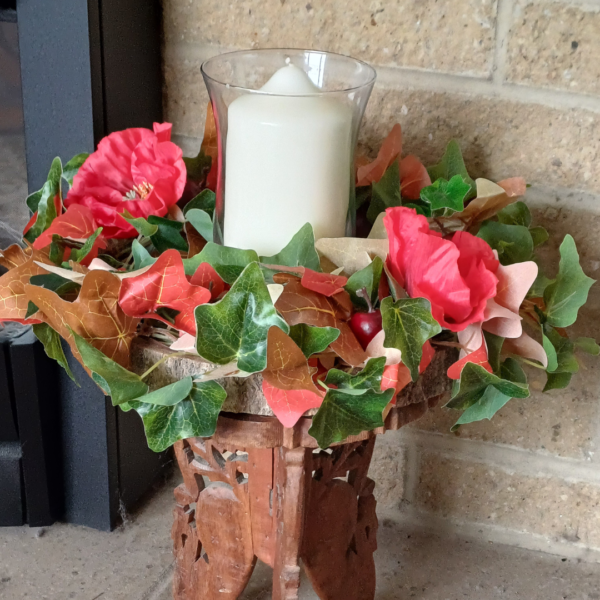 Image of table centre with green, read and brown ivy leaves, with two large red silk poppies. There is a large white pillar candle in the middle