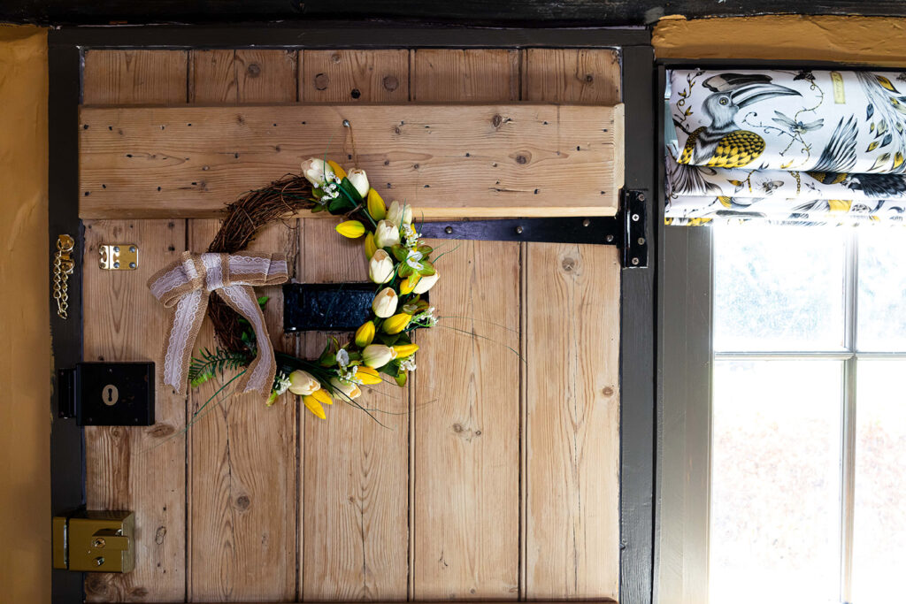 Image of tulip wreath on the inside of a rustic wooden door.  The tulips are yellow and white on a rustic grapevine wreath form, finished with a lacy hessian bow.