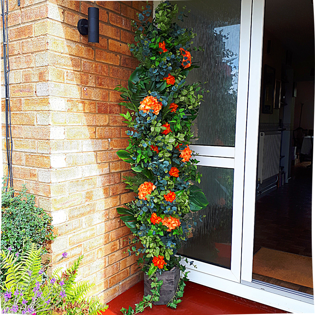 Picture of tall silk flower pillar with lush faux greenery and silk orange flowers. The pillar is installed as a feature in a corner of a plain front porch