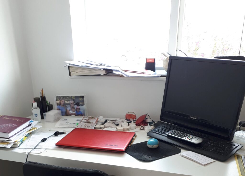Picture of home office desk, facing a window, with laptop and PC, and clutter of papers and pens