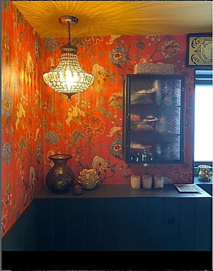 Image of part of a small bathroom.  Bold floral orange wallpaper, a crystal chandelier hanging lamp, quirky vases and ornaments on a black counter.  Bathroom accessories are hidden in a cabinet with smoky glass.