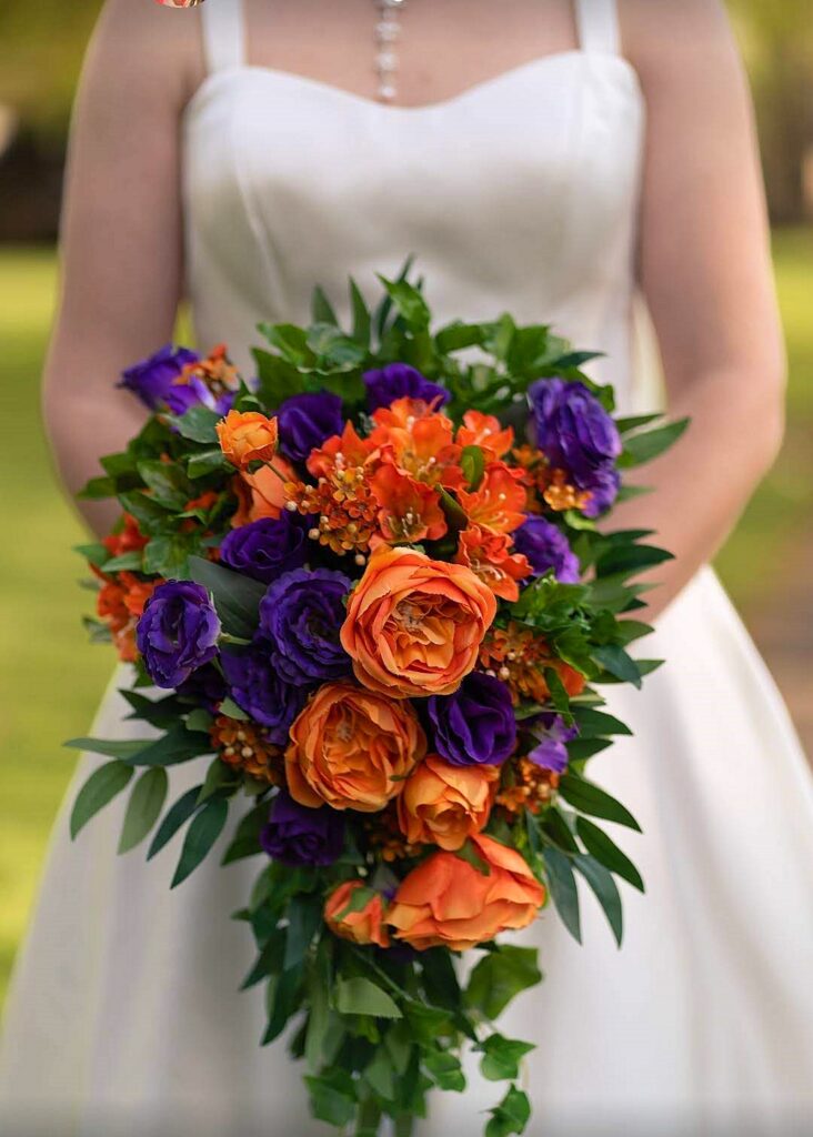 Image of cascade bouquet for bride;  The flowers are a mix of orange and purple blooms.  Roses, alstroemeria, lysianthus, ivy and willow