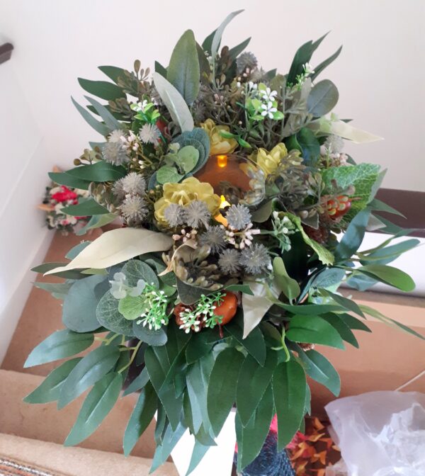 Image of table centre with green foliage, thistles, faux acorns and natural pinecones. A tealight flickers in the centre