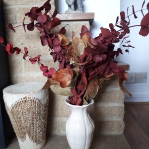 Image of a cream ceramic vase in a home hearth setting, with stems of red preserved eucalyptus.