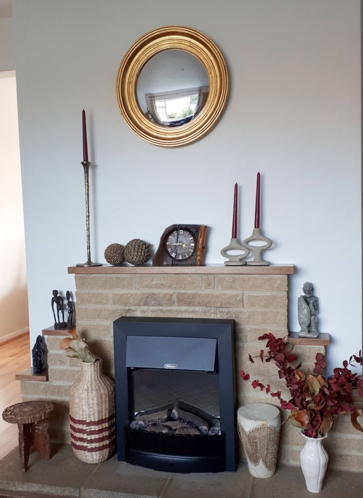 Style your home for autumn impact. Image of a fire place and mantelpiece. A round gold rimmed convex mirror is on the chimney breast. The mantel has candlesticks with burgundy candles, a wooden clock, and two bronze pinecones. Around the fireplace there is a cream ceramic vase with red preserved eucalyptus, a traditional African drum, a basket vase, and a smal carved stool.