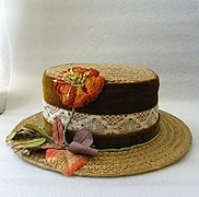 Image of a lady's straw hat with brown velvet round the crown, and a strip of lace over the velvet.  the hat is decorated with hand made orange silk flowers