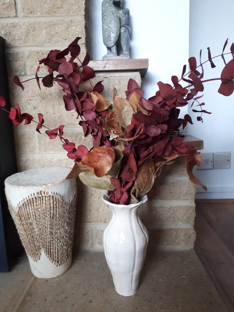 Image of red preserved eucalyptus in a cream ceramic vase. Next to the vase is an African traditional drum. On a little shelf above the vase is a soapstone carving of an African warrior