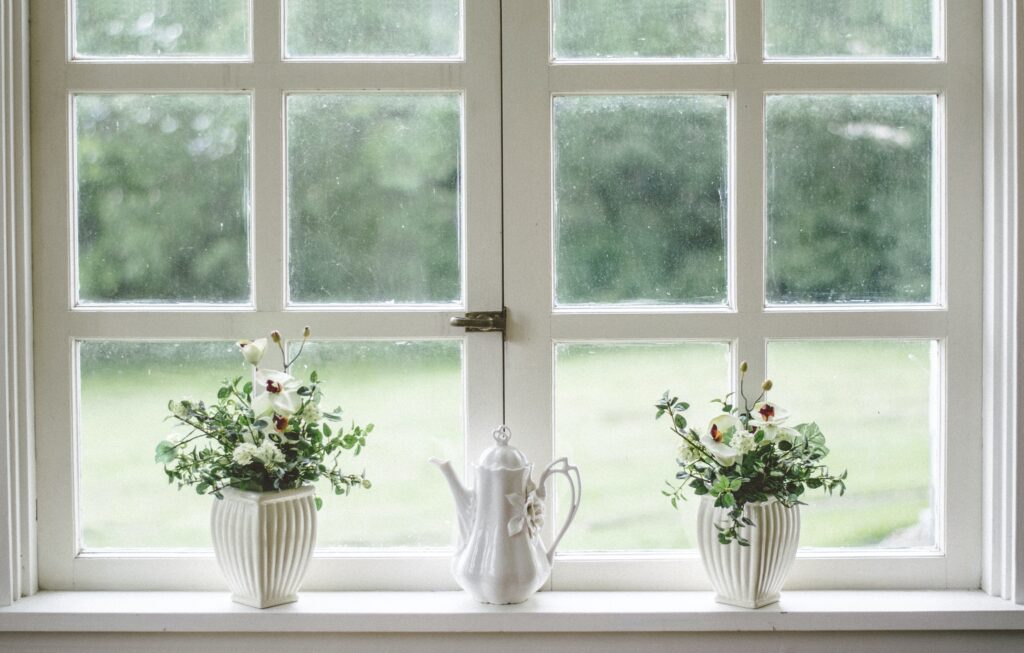 Two white vases with green and white arrangements on a windowsill, with an old fashioned white ceramic coffee pot in the middle.  The vases have a ribbed design