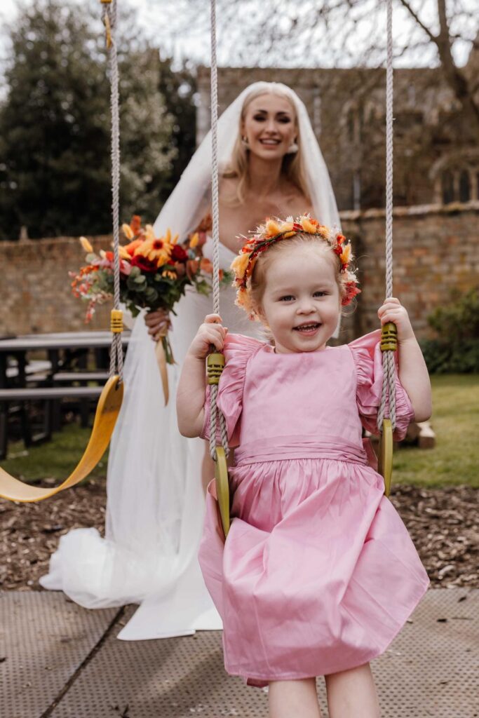 Flowergirl model with floral crown with red and ornage colours.  The little girl's dress is pink taffeta. In the background is the bridal model, with her sunset bridal bouquet
