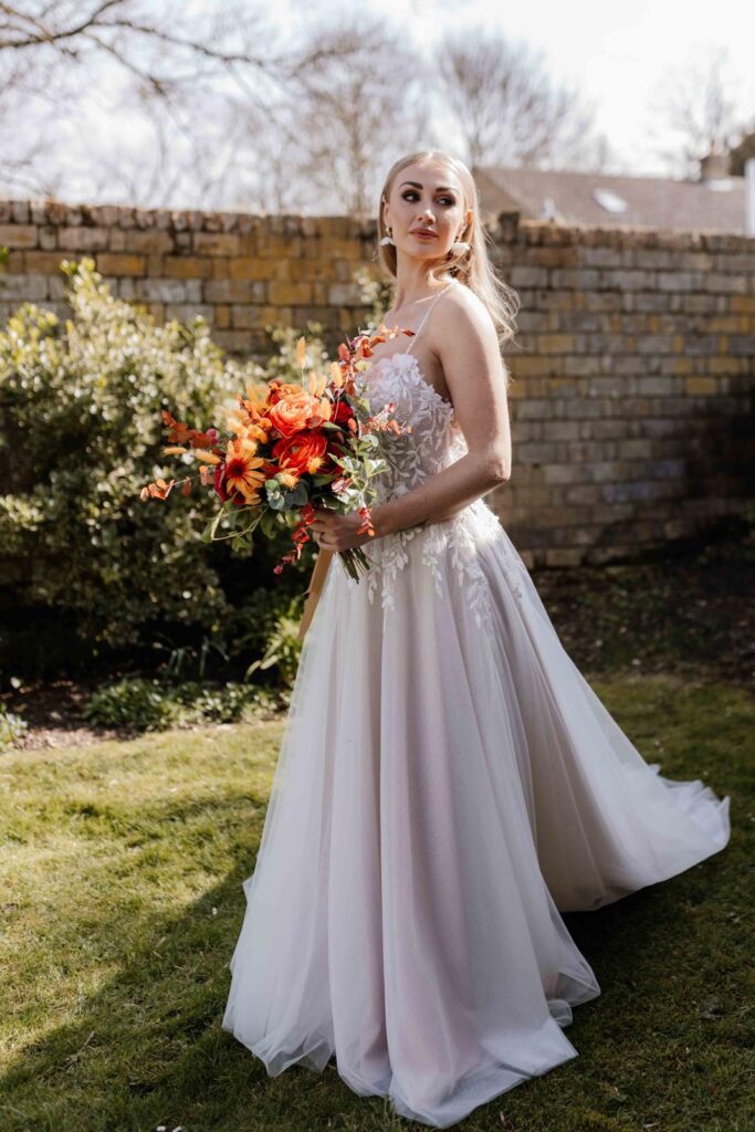 Model bride in lace and chiffon long dress, psed with bouquet of silk flowers with vibrant colours - orange, red, burgundy and green