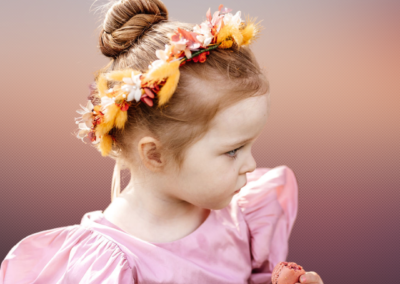 Image of little girl with a hair bun, and a floral crown made of silk flowers in deep orange and yellow, with orange bunnytail grasses