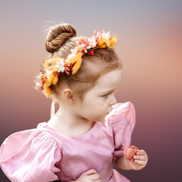 Image of little girl with a hair bun, and a floral crown made of silk flowers in deep orange and yellow, with orange bunnytail grasses