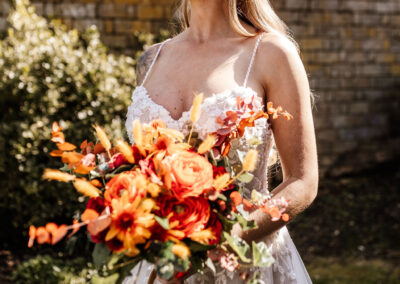 Image of bouquet of silk flowers in deep reds, orange and yellow. The bouquet is carried by a bride model