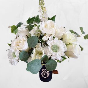 Image of white silk roses and other white flowers with green faux foliage in a dark blue vase. The vase has an ampisand carved with leafy flourishes