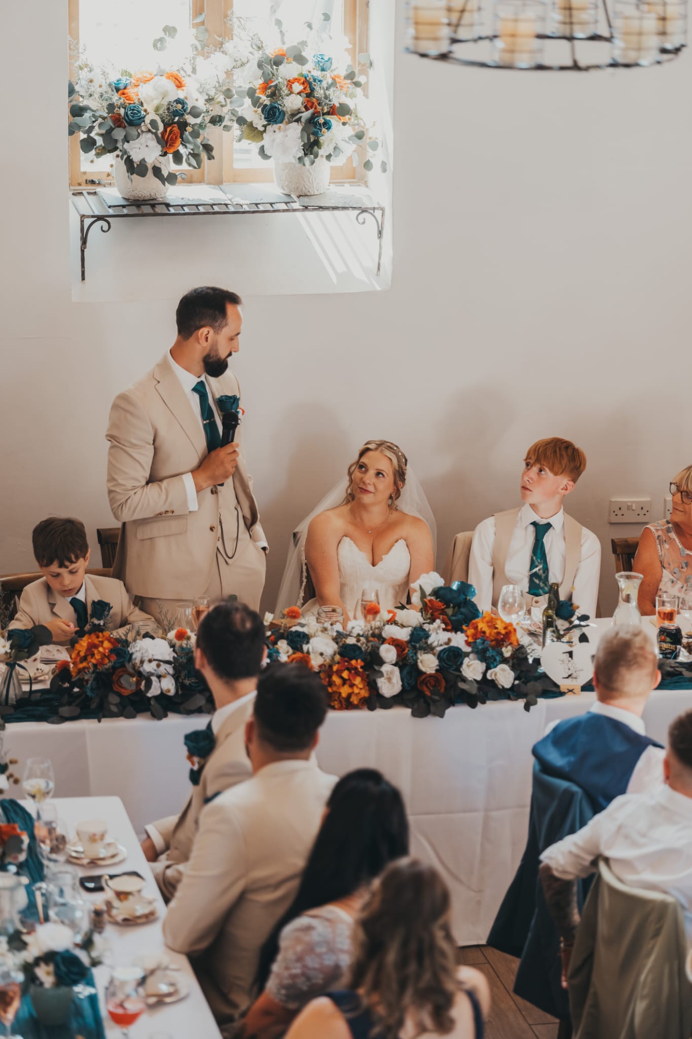 The Groom's speech to his bride. Bowls of flowers on the windowsil behind him, table decor with teal and terracotta flowers